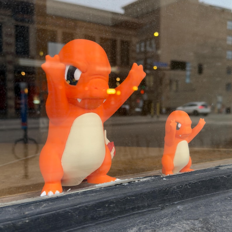 Puede incluir: Dos figuras de Charmander naranjas, una grande y otra peque&ntilde;a, est&aacute;n una al lado de la otra. La figura m&aacute;s grande tiene los brazos levantados, mientras que la m&aacute;s peque&ntilde;a imita la pose. Ambas tienen vientres color crema y se exhiben en una ventana.