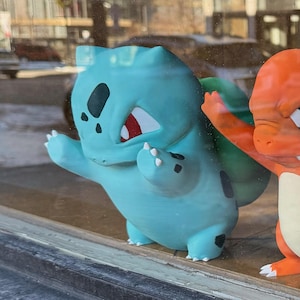 以下が含まれることがあります： 黒い斑点と赤いアクセントのある水色のフシギダネのフィギュア。背中には緑色の電球があります。隣にはオレンジ色のヒトカゲのフィギュアがあります。どちらも窓の前に立っています。