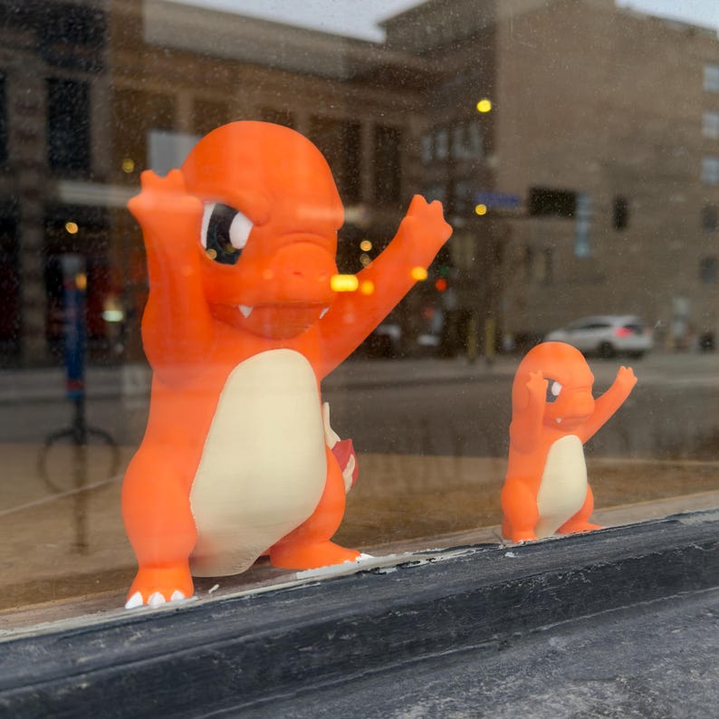 Puede incluir: Dos figuras de Charmander naranjas, una grande y otra peque&ntilde;a, est&aacute;n una al lado de la otra. La figura m&aacute;s grande tiene los brazos levantados, mientras que la m&aacute;s peque&ntilde;a imita la pose. Ambas tienen vientres color crema y est&aacute;n en un escaparate.