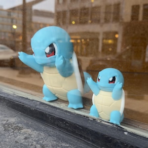 Puede incluir: Se muestran dos figuras azules de Squirtle, una grande y otra peque&ntilde;a. Las figuras tienen cuerpos azul claro, caparazones beige y detalles rojos alrededor de los ojos. Posan con los brazos levantados, dando una apariencia juguetona.