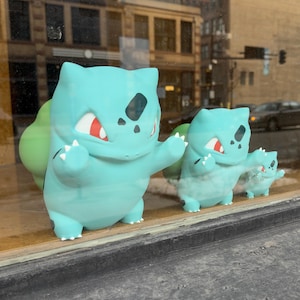 Puede incluir: Se exhiben tres figuras de Bulbasaur de color azul claro de diferentes tama&ntilde;os. Las figuras tienen ojos rojos, garras blancas y un bulbo verde en la espalda. Est&aacute;n colocadas frente a una ventana, con una calle de la ciudad visible al fondo.