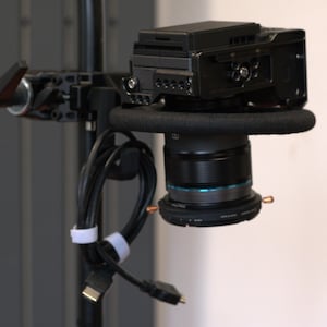 May include: A black digital camera mounted on a black rig with a lens and attached cables. The camera is positioned on a padded support, with the lens featuring a blue accent ring. The cables are secured with white fasteners.