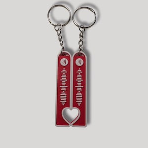 May include: Two keyrings with red rectangular pendants featuring a white heart-shaped cutout and a Spotify code design. Each keyring has a silver chain and a silver key ring. The pendants are connected to form a heart.