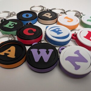 May include: A selection of alphabet keyrings in various colours. Each keyring has a circular design with a letter in a contrasting colour against a black or white background. The keyrings have silver key rings.
