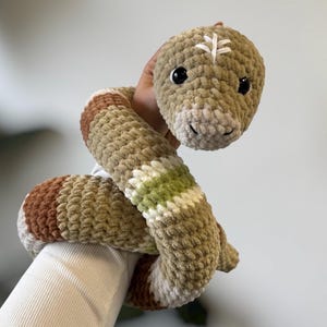 Könnte beinhalten: Ein gehäkeltes Schlangenspielzeug in Beige-, Braun- und Grüntönen. Die Plüschschlange hat ein lächelndes Gesicht mit schwarzen Augen und ein weißes, aufgesticktes Design auf dem Kopf. Der Körper hat gestreifte Farbbereiche.