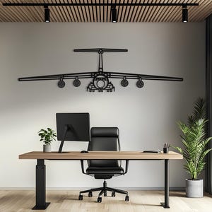 May include: A black metal wall art piece depicting the silhouette of a large cargo aircraft with a T-tail design, four under-wing engines, and a prominent fuselage, mounted horizontally on a plain wall above a desk and office chair.