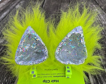 Pode incluir: Um par de prendedores de cabelo verde limão felpudo com orelhas prateadas iridescentes. A embalagem inclui instruções para limpeza a seco e lavagem em uma bolsa de malha. A embalagem também indica que os prendedores são para crianças de 3 anos ou mais.