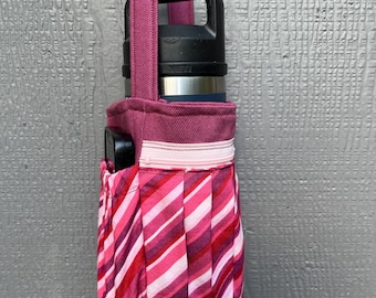 Pode incluir: Uma bolsa de tecido de rayas rosa e brancas com fecho de correr. A bolsa tem uma alça rosa e contém uma garrafa de água preta e prateada.