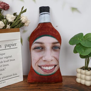 May include: A novelty cushion in the shape of a ketchup bottle, with a smiling face printed on it. The bottle is red with a green band and a black cap. The face has blue eyes and a wide smile. The cushion is on a wooden surface.