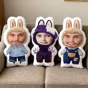 Peut inclure: Trois coussins personnalisés en forme de lapin avec des visages imprimés. Les coussins présentent différentes tenues, dont une salopette en jean bleu, une chemise rayée bleue et une tenue à capuche violette. Les coussins sont sur un canapé beige.
