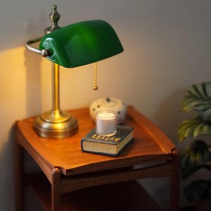 May include: Miniature wooden side table with a green-shaded brass lamp, a small candle, and a book. The lamp has a pull chain. The book has gold lettering and a blue cover. A small white and blue decorative item sits behind the candle.