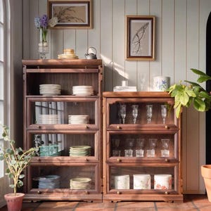 May include: Two wooden display cabinets with glass doors, filled with stacks of white and patterned plates, glasses, and decorative items. The cabinets are set against a light-colored wall with framed artwork. The text "ME&HOUSE" is visible.