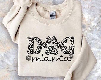 May include: A beige sweatshirt with the words "DOG mama" in black script. The letters "DOG" are designed with a leopard print pattern, and a paw print graphic is in the center. Two small paw prints are on either side of the word "mama".