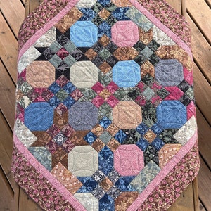 May include: A quilt with a geometric pattern in shades of brown, pink, blue, and grey. The quilt is made of fabric with floral and paisley designs. The quilt is bordered with a pink and brown floral fabric.
