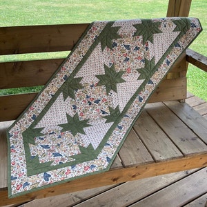 May include: A green and white table runner with a starburst pattern. The runner is made of fabric with a floral print and is laid out on a wooden bench.