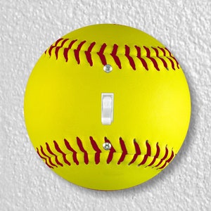 May include: A yellow softball light switch cover with red stitching. The switch is white and is in the center of the softball.