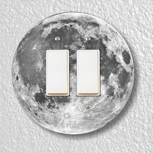 May include: A round light switch cover with a black and white image of the moon. The cover has two white toggle switches.
