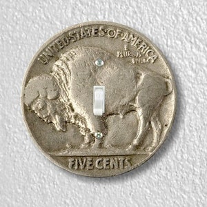 May include: A silver-colored US five-cent coin with a buffalo design and the text "UNITED STATES OF AMERICA" and "FIVE CENTS" on it. The coin has a light switch in the center.