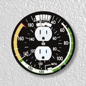 May include: A black and white gauge with a yellow and green arc. The gauge has a needle pointing to 40 and two electrical outlets on the face. The gauge is labeled "M.P.H."