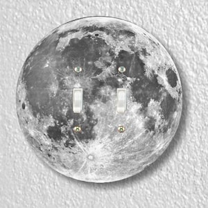 May include: A round light switch cover with a black and white image of the moon. The switch cover has two toggle switches.