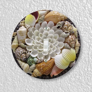 May include: A round light switch cover with a decorative design of seashells. The cover features a variety of seashells in different shapes, sizes, and colors, including white, brown, and yellow. The seashells are arranged in a circular pattern around a white light switch.