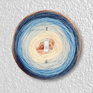 May include: A round light switch cover with a blue, white, and peach yarn design. The yarn is wound in a circular pattern, creating a gradient effect.