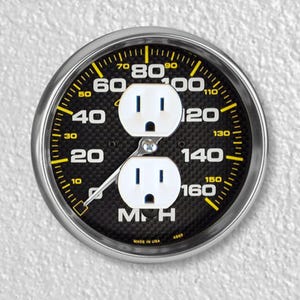 May include: A black and yellow speedometer with a carbon fiber design. The speedometer has two electrical outlets in place of the needle. The speedometer reads "MPH" and "Made in USA".
