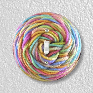 May include: A round light switch cover with a colorful yarn design. The yarn is in shades of pink, blue, green, yellow, and orange. The switch is white and is in the center of the cover.