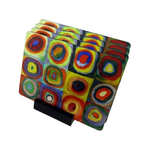 May include: Set of four colorful glass coasters featuring a geometric pattern of squares and circles in shades of red, yellow, blue, green, and purple. The coasters are stacked on a black stand.