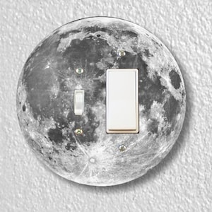 May include: A round light switch cover with a black and white image of the moon. The switch cover has a white light switch with a silver toggle.
