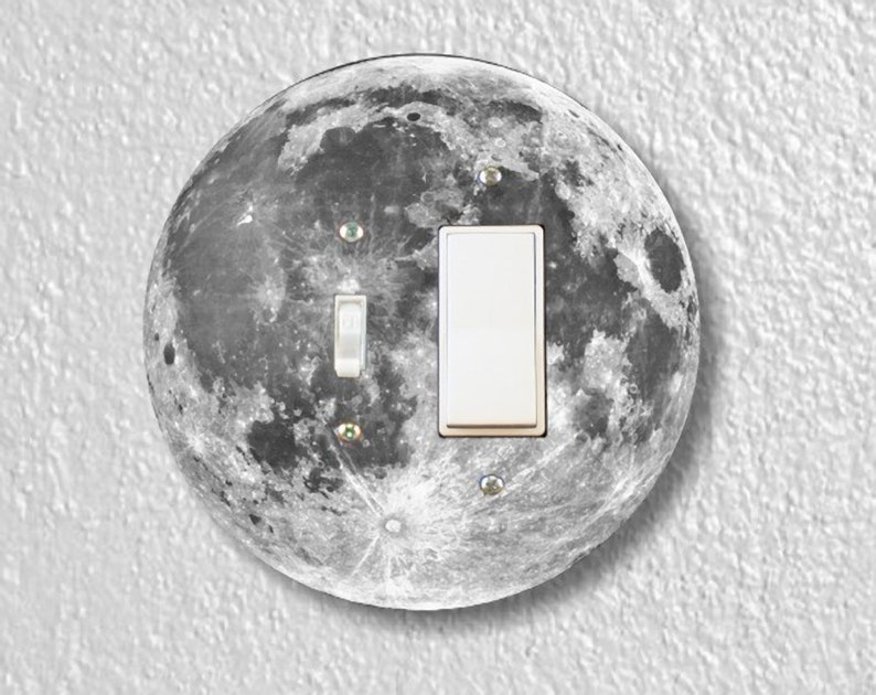 May include: A round light switch cover with a black and white image of the moon. The switch cover has a white light switch with a silver toggle.
