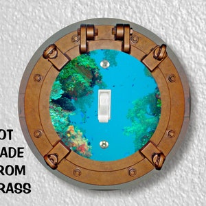 May include: A round light switch cover with a nautical porthole design. The porthole is open and shows a blue underwater scene with coral and fish. The switch cover is made of plastic and has a brown finish.