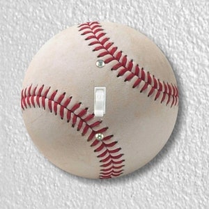 May include: A baseball-themed light switch cover. The cover is designed to look like a baseball, with red stitching and a white surface. The switch is white and the screws are silver.