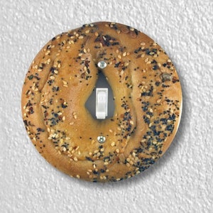 May include: A light switch cover with a photo of a round, brown everything bagel with poppy seeds, sesame seeds, and black sesame seeds.