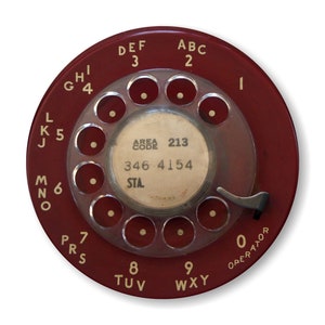May include: A red rotary dial phone with a white dial and black lettering. The dial has the letters ABC, DEF, etc. around the outside and the numbers 1 through 0 in the center. The dial also has the text "AREA CODE 213 346 4154 STA."