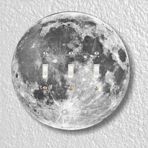 May include: A light switch cover featuring a black and white image of the moon with three white toggle switches and three small green eyes.