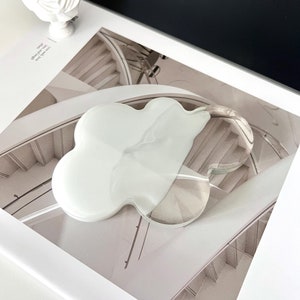 May include: A cloud-shaped, clear acrylic coaster with a white, marbled design. The coaster sits on a photograph of a staircase. A white bust of a classical figure is in the background.