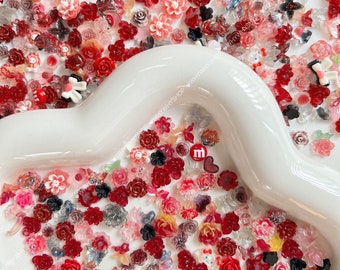 May include: A collection of small, decorative floral embellishments in shades of red, pink, and clear. The assortment includes roses, other flower shapes, and some with butterfly and heart designs. The items are scattered on a white, curved surface.