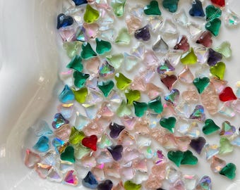 May include: A white ceramic dish filled with a variety of small, heart-shaped glass rhinestones. The rhinestones are in shades of green, blue, red, pink, and clear.