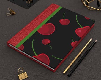 May include: A closed journal with a red textured cover and a black cover featuring a pattern of red cherries with green stems. A green stripe accents the spine. Gold and black pencils and a gold binder clip are nearby.