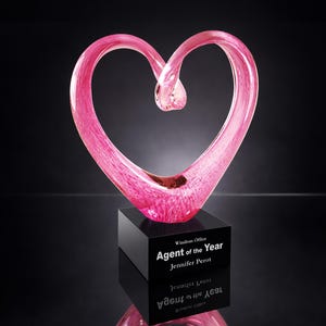 May include: A pink glass heart-shaped award on a black base. The award has the text "Agent of the Year" and "Jennifer Perot". The heart is a vibrant pink color and sits on a reflective surface.