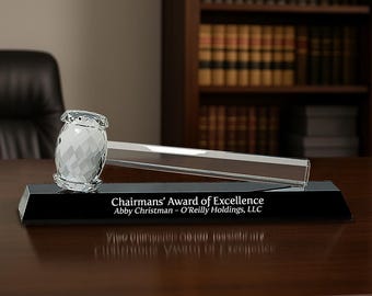 May include: A clear crystal gavel with a faceted head rests on a rectangular black glass base. The base features the engraved white text: Chairmans' Award of Excellence, Abby Christman - O'Reilly Holdings, LLC, centered on the front face.