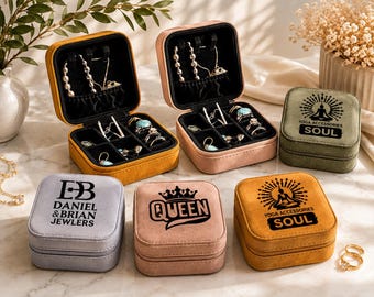 May include: A collection of six square velvet jewelry boxes in mustard yellow, dusty pink, olive green, and light gray. Two boxes are open, revealing black velvet interiors with necklace hooks and ring slots. Others are closed with printed logos.