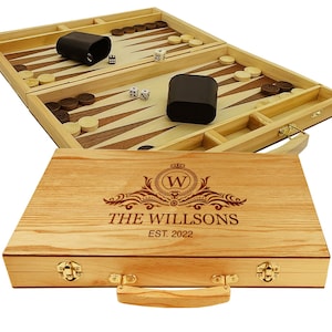 May include: A wooden backgammon set with a personalized inscription that reads "The Willson's Est. 2022". The set features a wooden case with a handle and brass clasps. The backgammon board is open and shows the playing surface with checkers and dice.