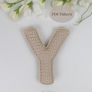 May include: A crocheted letter "Y" in a neutral beige color, lying on a white surface. The handmade letter is surrounded by white flowers and greenery. The text "PDF Pattern" is visible in the background.