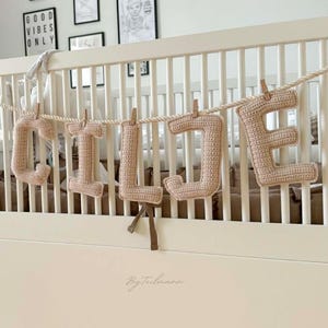 Może przedstawiać: Beżowy szydełkowy baner z imieniem "LILIE" zawieszony nad białym łóżeczkiem dziecięcym. Litery przymocowane są do sznurka za pomocą małych drewnianych spinaczy. Łóżeczko stoi w pokoju z oprawionymi grafikami i napisem "GOOD VIBES ONLY".