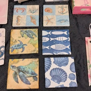 May include: A selection of square coasters and decorative items with a coastal theme. Designs include a flamingo, octopus, sea turtles, fish, and seashells. The colour palette is primarily pink, blue, and beige. A pink banner with a flamingo and a pink heart-shaped decoration are also visible.