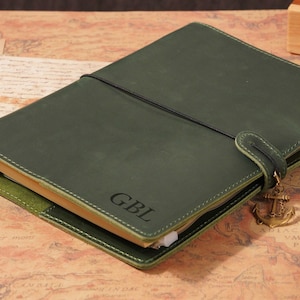 Peut inclure: Journal en cuir vert avec les initiales "GBL" estampées sur la couverture. Le journal est sécurisé par un élastique noir et une sangle en cuir avec un charme d'ancre dorée. Le journal est ouvert sur une carte.
