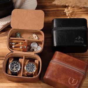 Può includere: Un portagioie da viaggio in pelle marrone aperto con gioielli e due orologi. Sono presenti anche due custodie per orologi in pelle nera e un astuccio in pelle marrone con cerniera. La custodia nera ha il nome "Audrey" stampato.