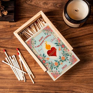 Puede incluir: Caja de cerillas de madera con cerillas y una tarjeta decorativa. La tarjeta presenta un corazón con una llama, detalles florales y el texto "Faites l'un pour l'autre" y el año 2025. También se ve una vela encendida.